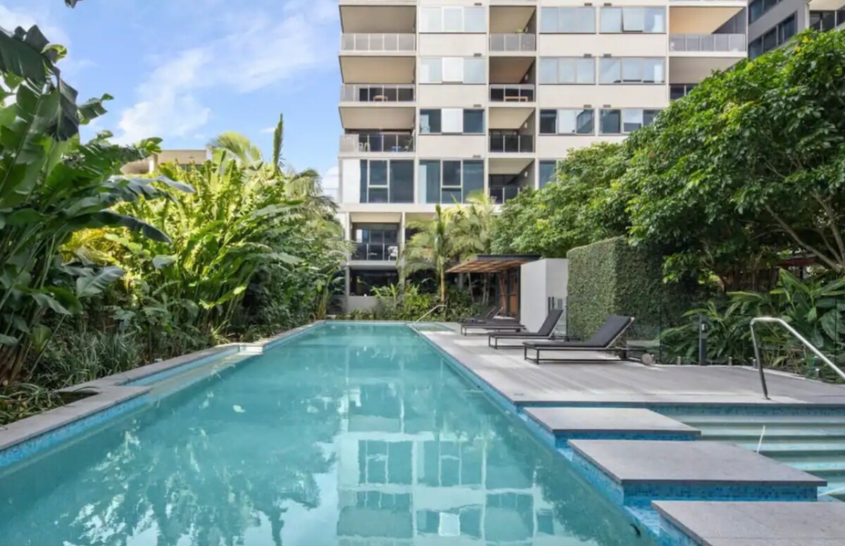 A serene outdoor pool is surrounded by lush greenery and modern lounge chairs. The clear water reflects the surrounding plants and the building's contemporary architecture, creating a peaceful retreat for relaxation.