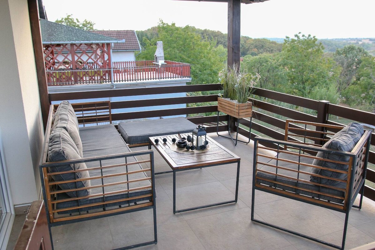 A spacious balcony area features comfortable seating arranged around a low table. Natural greenery is visible in the background, enhancing the outdoor space. Potted plants add a touch of nature, creating a serene environment for relaxation.