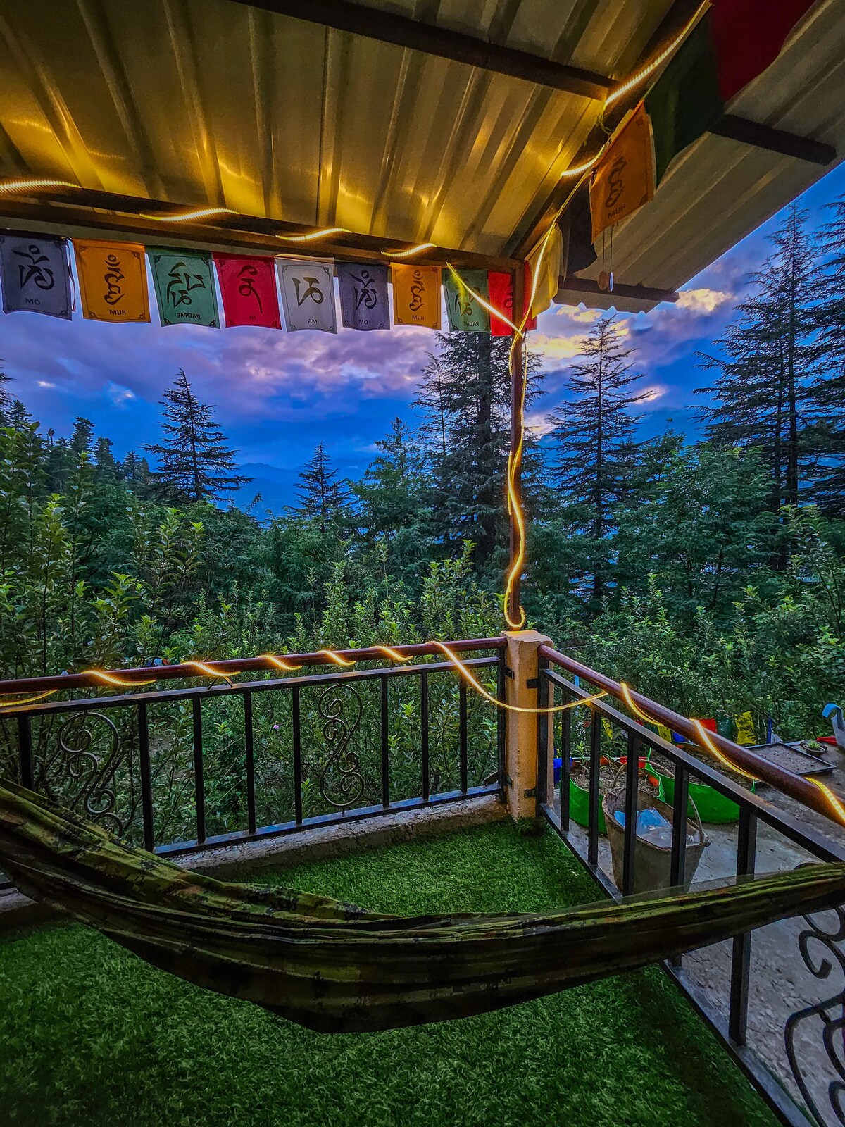 A balcony features a hammock alongside vibrant prayer flags, with lush greenery and distant mountains visible. Soft lighting enhances the space, creating an inviting environment for relaxation. The view captures the serene ambiance of nature, complemented by a colorful sky at dusk.