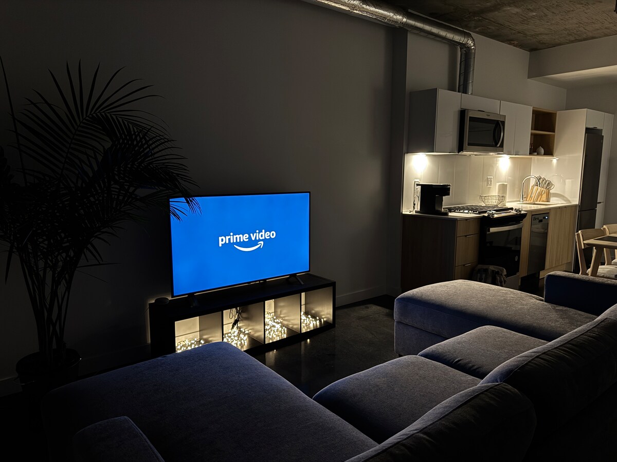 A modern living area features a large sectional couch opposite a sleek television displaying the Prime Video logo. Soft, ambient lighting illuminates a coffee table adorned with decorative elements, while a plant adds a touch of greenery to the space.
