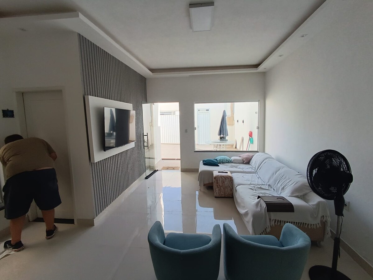 A bright living area features a comfortable sofa, a modern flat-screen TV mounted on the wall, and large windows allowing natural light. A small table and two armchairs are positioned near the windows, enhancing the spacious feel of the room.