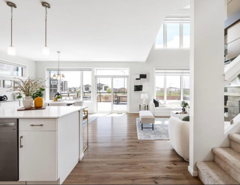 A bright and open living space is seen featuring large windows allowing natural light. A light-colored kitchen with modern fixtures overlooks a cozy sitting area with a comfortable sofa. Stairs are visible in the background, leading to an upper level.