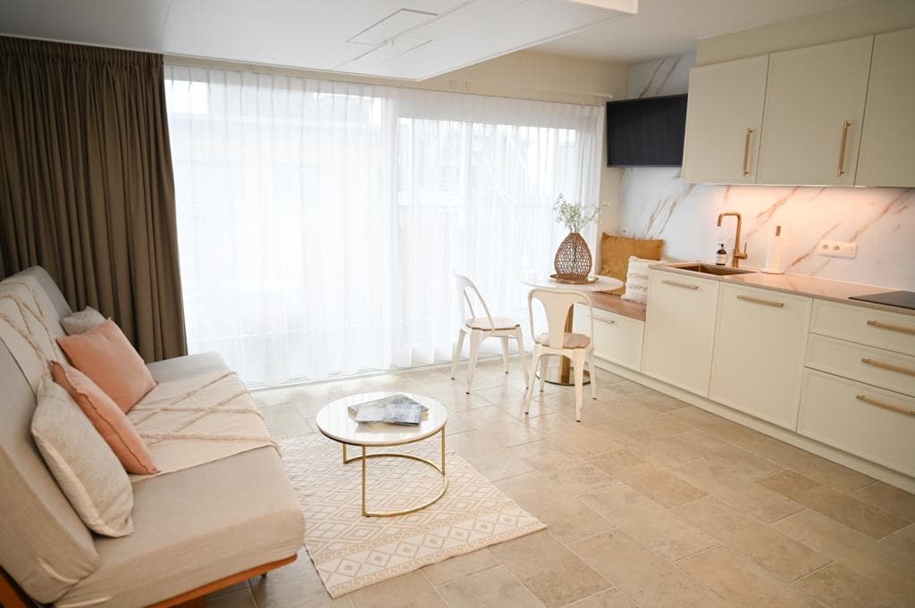 An inviting living space features a light-colored sofa adorned with decorative pillows, accompanied by a circular coffee table. Natural light filters through sheer curtains, illuminating the kitchen area with modern cabinetry and a small dining table, enhancing the cozy atmosphere.