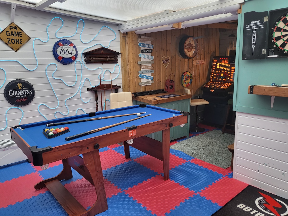 A games room is equipped with a blue pool table, a dartboard, and a vintage-style gaming machine. Colorful floor tiles create a playful atmosphere. The wooden walls are decorated with various signs, adding character to the inviting space.