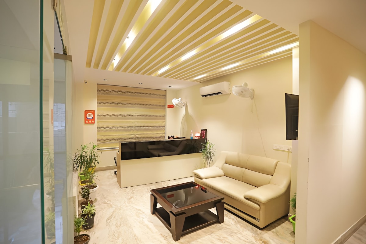 A modern reception area features a light-colored sofa and a dark coffee table. A sleek front desk is positioned against the back wall, with decorative plants enhancing the space. Soft overhead lighting creates a welcoming ambiance within the room.