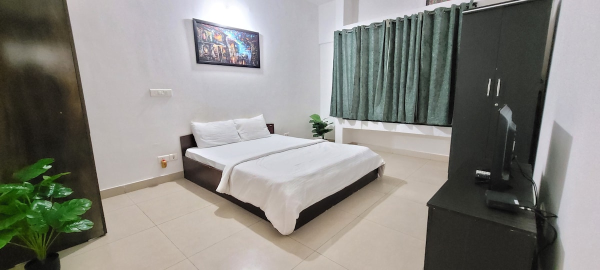 A clean and spacious sleeping area features a large bed with a white comforter. A black cabinet and a television stand are present, along with a small green plant. Natural light enters through the window, which is covered by dark green curtains.