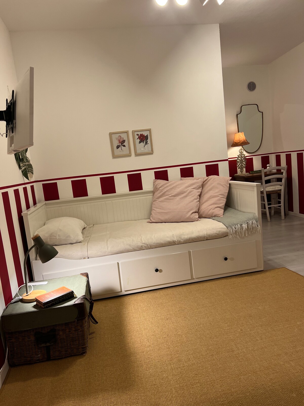 A cozy interior space features a daybed adorned with soft pink cushions, set against a backdrop of red-striped walls. A small side table with a lamp provides gentle lighting, while a mirror adds depth to the room. A woven area rug covers the floor.