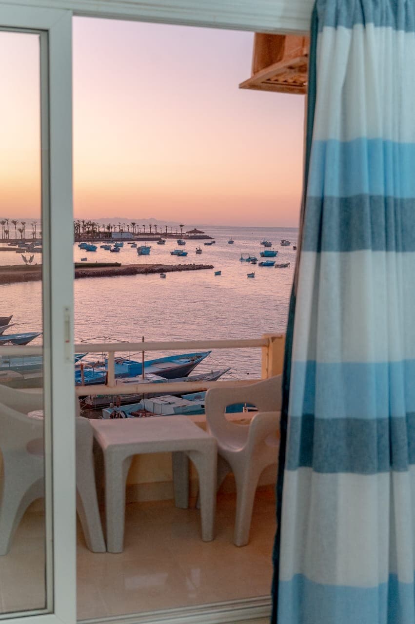 A view of the sea is framed by sheer curtains with blue stripes. Two white chairs are positioned on the balcony, overlooking boats scattered across the water at sunset, creating a serene atmosphere.
