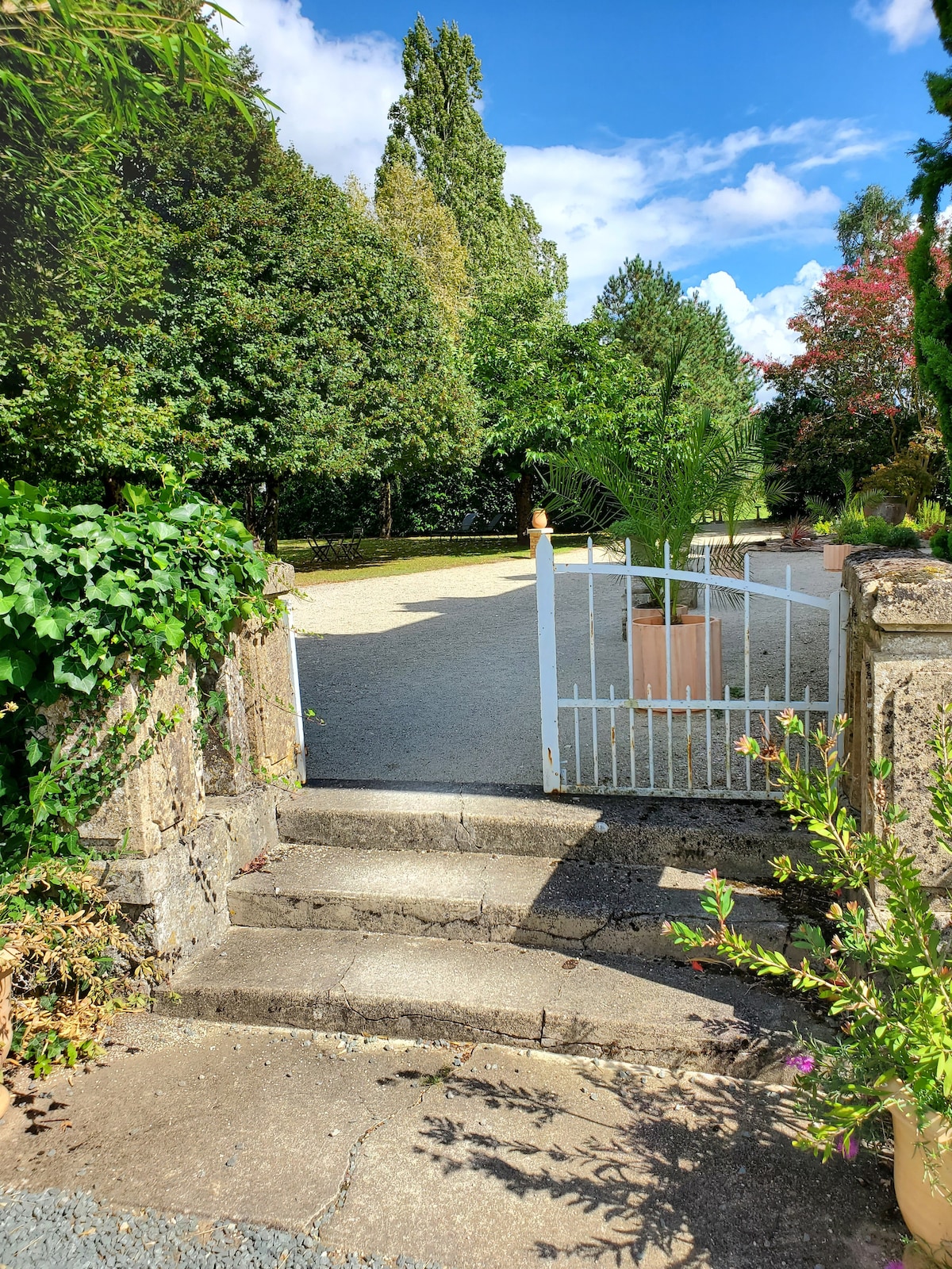 An entrance gate is visible, framed by leafy greenery and potted plants. A gravel pathway leads into a spacious outdoor area surrounded by trees. Steps made of stone provide access to the gate, while sunlight brightens the scene, highlighting the inviting landscape.