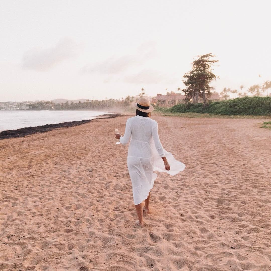 A sandy beach is seen at sunrise, with soft waves gently lapping at the shore. A person in a flowing white dress is walking along the beach, holding a glass. Lush greenery and palm trees are visible in the background, suggesting a tranquil setting.