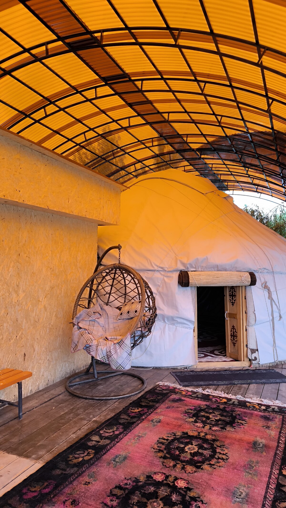 A yurt is positioned under a covered terrace, featuring a rounded exterior and an entrance adorned with decorative elements. A cozy hanging chair is placed nearby, accompanied by a patterned rug on the wooden floor, enhancing the warm and comfortable ambiance.