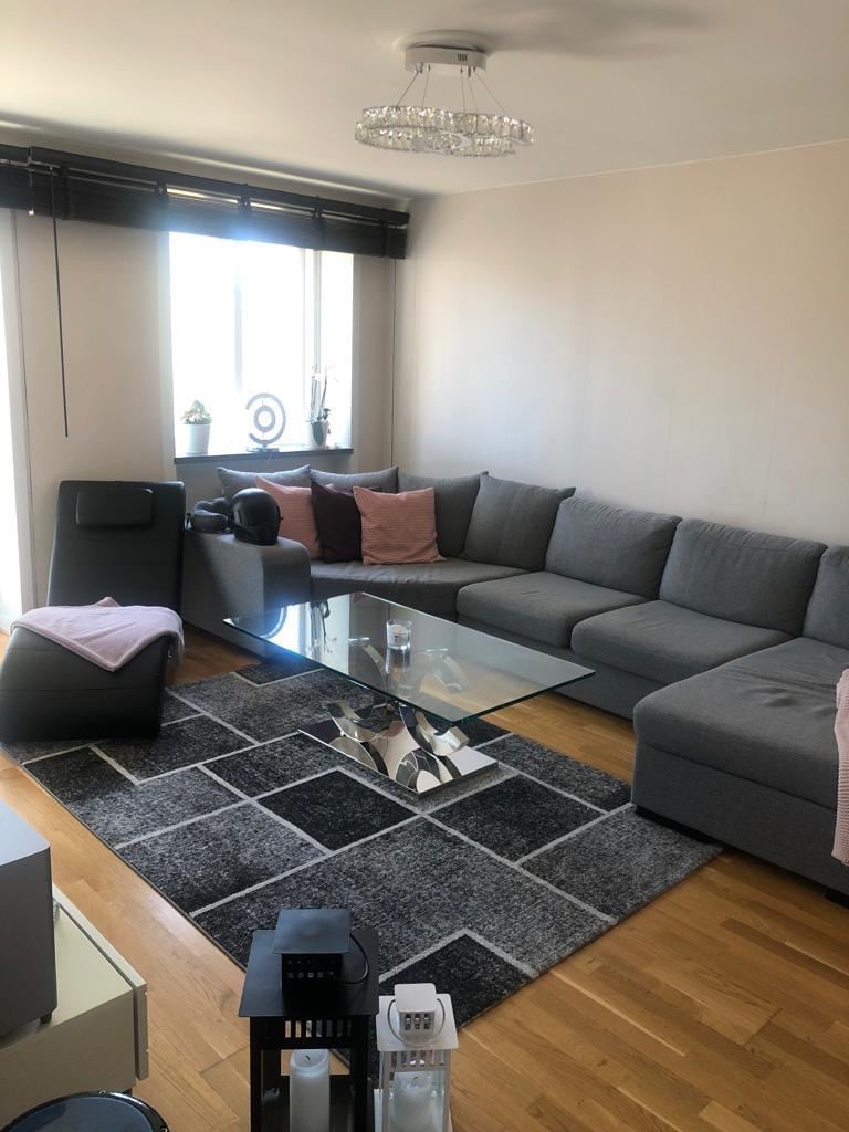 A modern living room features a large grey sectional sofa with pink accent cushions, arranged around a glass coffee table. A patterned rug lies on the wooden floor, providing a contrast with the light-coloured walls. Natural light enters through a window dressed with dark blinds.