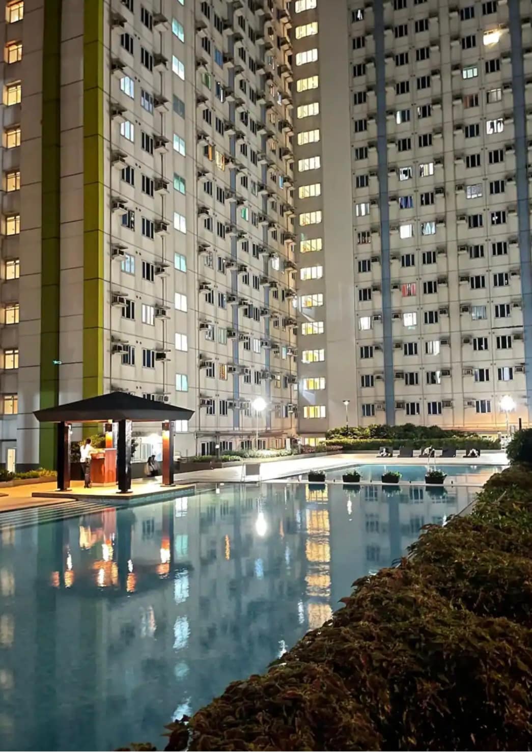 A tranquil pool is surrounded by well-lit residential buildings, with soft reflections shimmering on the water's surface. Lounge chairs are arranged neatly beside the pool, while a shaded gazebo offers a spot for relaxation amidst the serene evening ambiance.