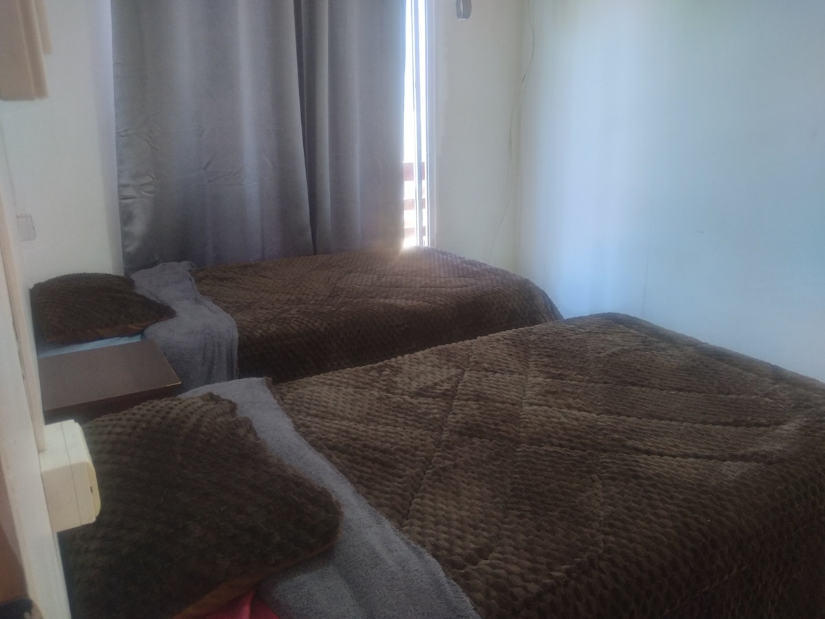 A cozy sleeping area is shown with two beds dressed in soft brown blankets. Light enters through a partially opened window, and grey curtains hang alongside the walls. Nightstands are placed beside each bed, adding to the room's comfortable layout.