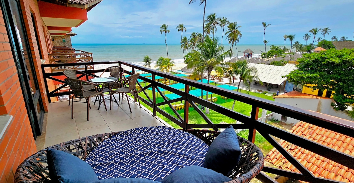 A spacious balcony features a round wicker chair with navy blue cushions, offering views of palm trees and the ocean. A small table with two chairs is positioned nearby, overlooking green gardens and multiple pools on the property.