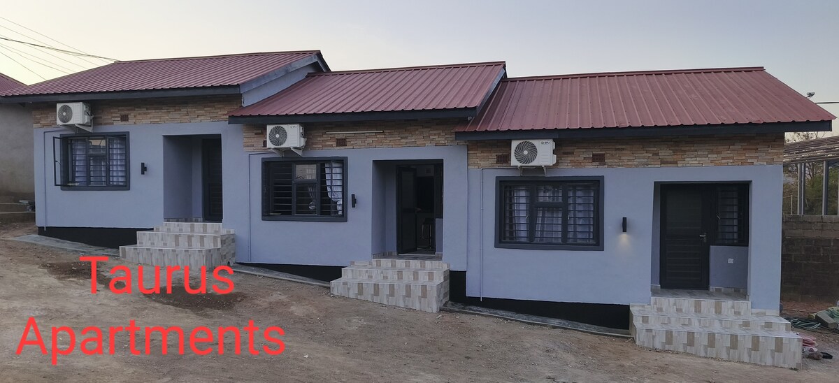 An exterior view of Taurus Apartments showcases a modern structure with a distinctive red metal roof. The building features air conditioning units and stone accents. A set of stairs leads to the entrances, which are framed by windows that allow for natural light.