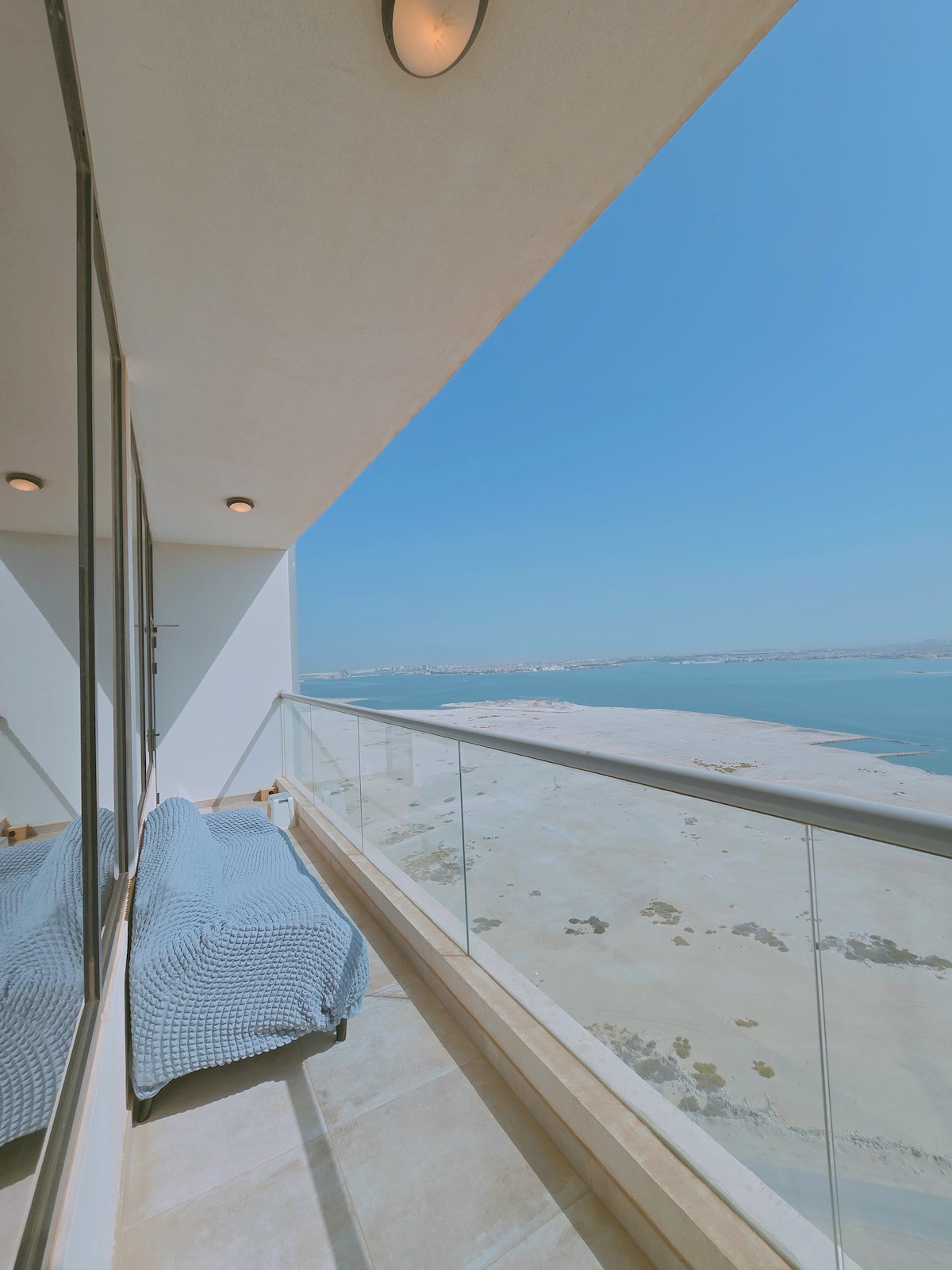 A private balcony offers expansive views of the sea and nearby landscape. The glass railing accentuates the openness, while comfy seating is visible. Natural light floods the area, reflecting the open sky and tranquil surroundings.