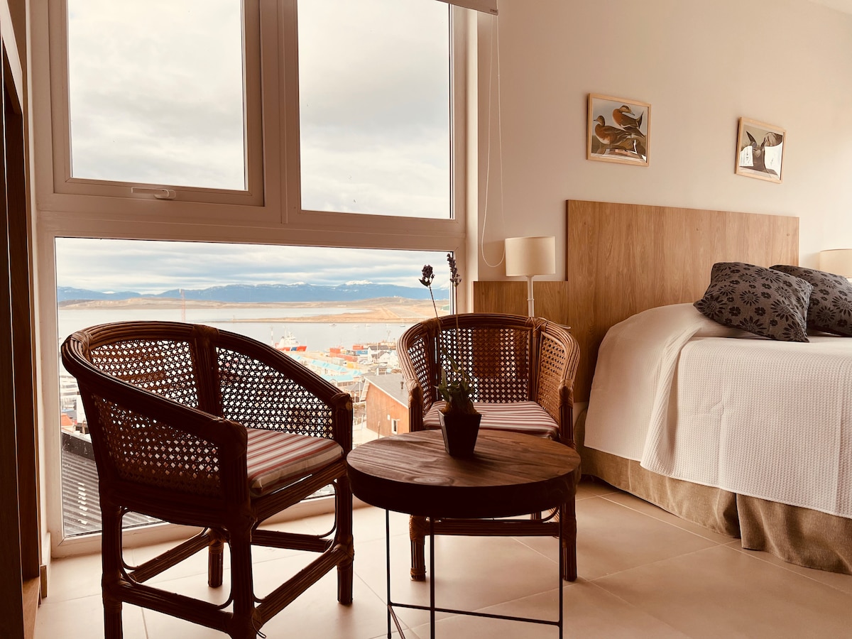 A cozy corner of a room features two woven chairs and a circular wooden table. A spacious bed with neutral linens is positioned nearby, and large windows offer a view of the outside landscape, allowing natural light to brighten the space.