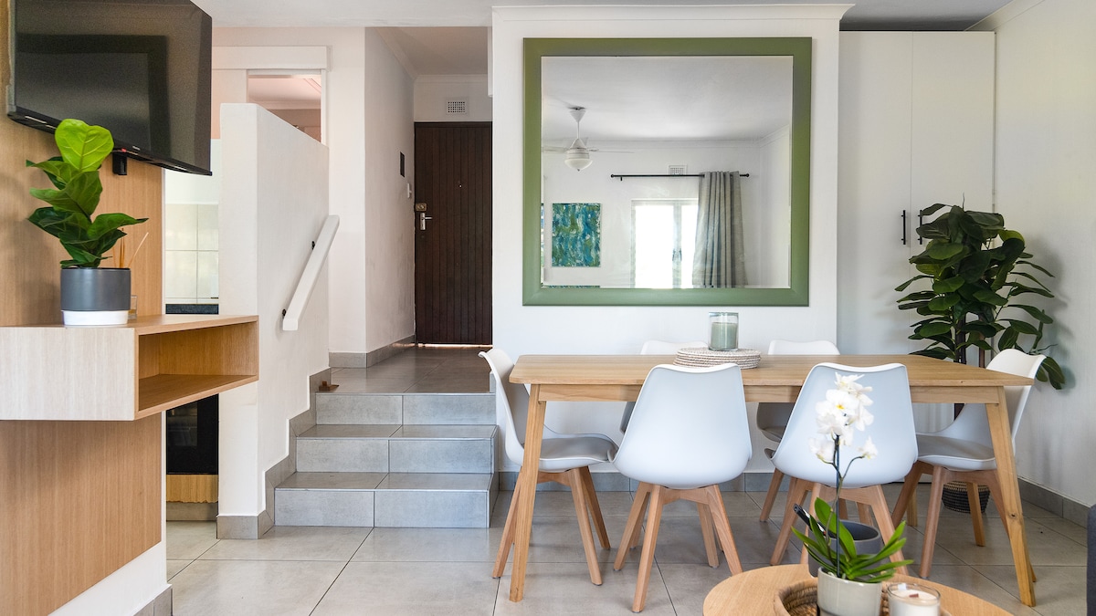 A bright dining area features a wooden table surrounded by modern white chairs. A large green framed mirror reflects the space, enhancing the natural light. Indoor plants are positioned on the table and nearby, while a television is mounted on the wall, providing entertainment options.