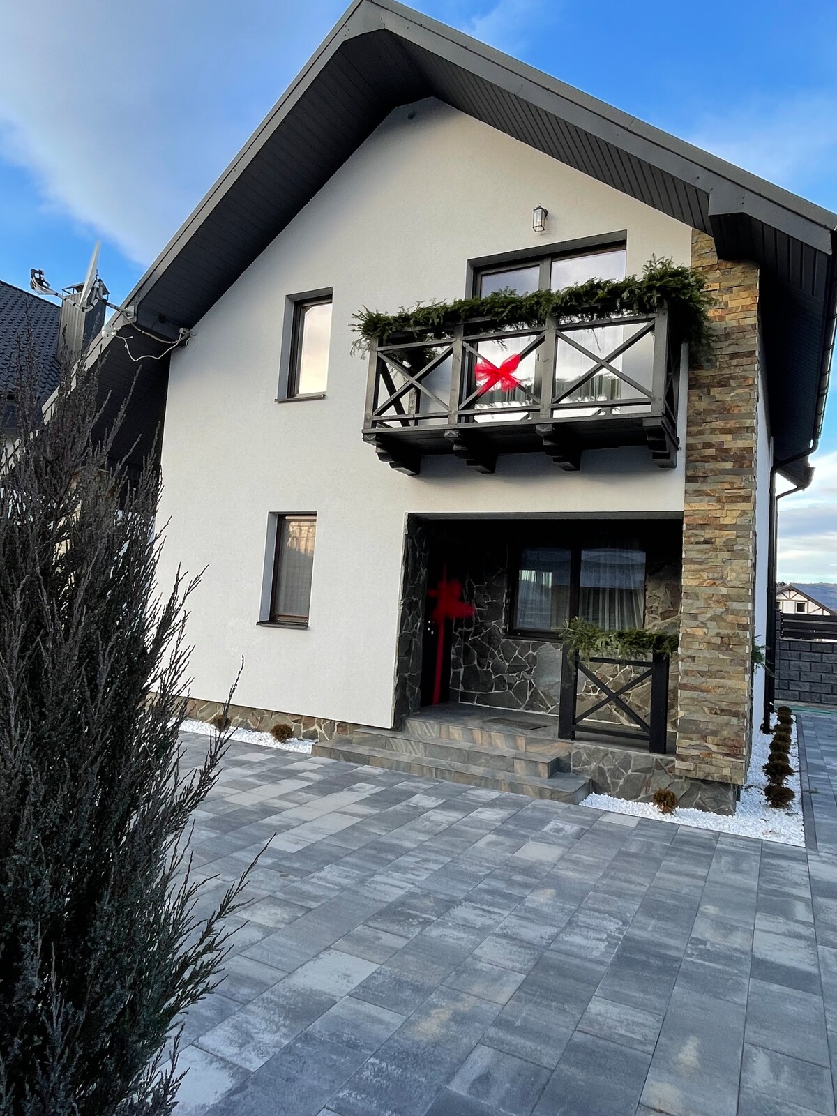 A modern home features a white exterior with a stone accent wall. A balcony adorned with greenery stretches across the upper floor, complemented by a red decorative element. The driveway is paved with grey stones, surrounded by carefully positioned shrubs and small trees.