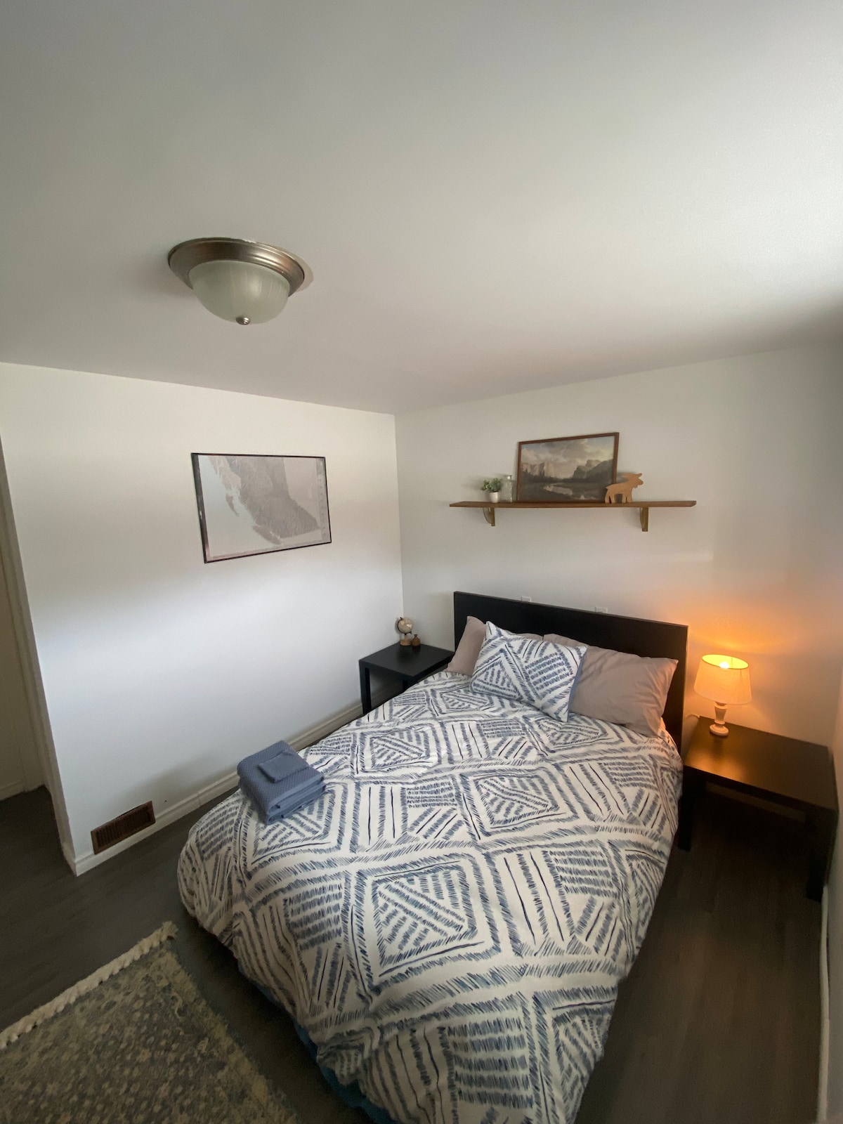 A cozy sleeping area features a spacious bed with a patterned duvet cover. Soft lighting from a bedside lamp creates a warm glow. A wall-mounted shelf holds decorative items, while a minimalist artwork hangs above the bed, contributing to a simple but inviting ambiance.