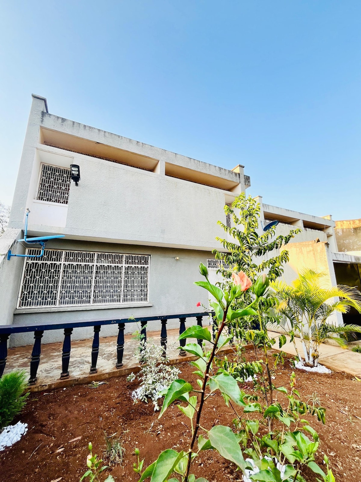 A two-story exterior of a retreat is visible, featuring a light-colored facade. Large windows with grills allow natural light to filter in. Tropical plants and flowers adorn the carefully landscaped garden, adding greenery to the scene under a clear blue sky.