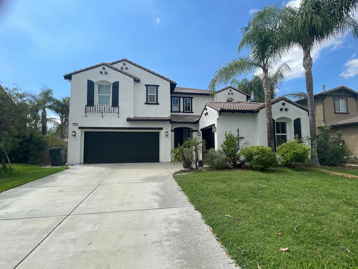 The home's exterior is characterized by a mix of white stucco and dark brown accents, featuring a tiled roof. Lush greenery surrounds the driveway, which leads up to a spacious two-car garage, while palm trees add to the inviting landscape.