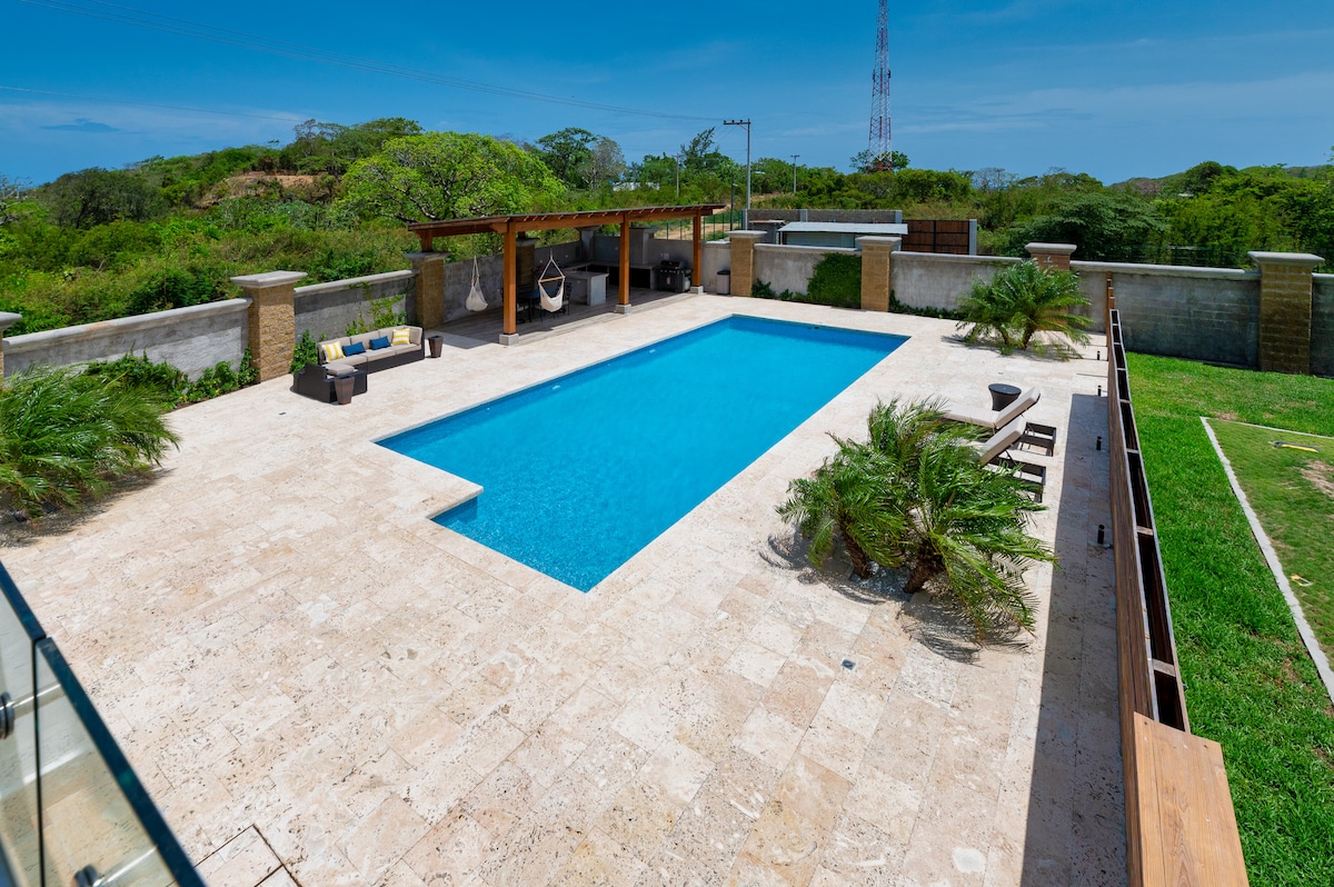 An outdoor area features a large swimming pool bordered by light-colored stone tiles. Lounge chairs are placed on one side, while a shaded seating area includes a hanging chair and a small table. Lush greenery surrounds the space, adding a natural touch.