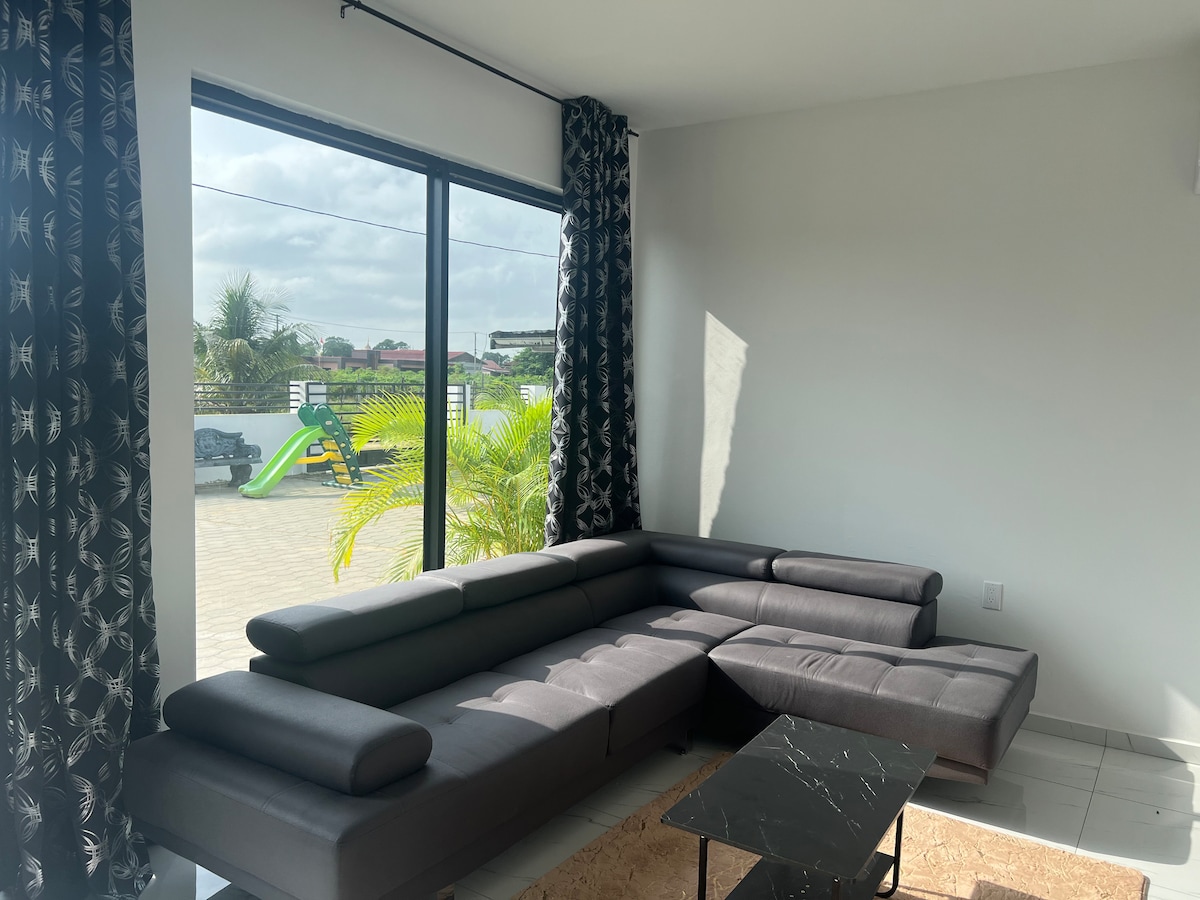 A modern living area features a spacious, sectional sofa positioned against a large glass window. Sunlight filters through patterned curtains, illuminating the space. A small black coffee table is situated on a textured rug, with an outdoor view of greenery and a play area beyond.