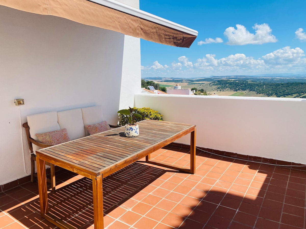 A sunlit terrace features a wooden dining table surrounded by comfortable seating. A potted plant sits at the center of the table. Scenic views of rolling hills and a clear blue sky are visible in the background, enhanced by a partial awning.