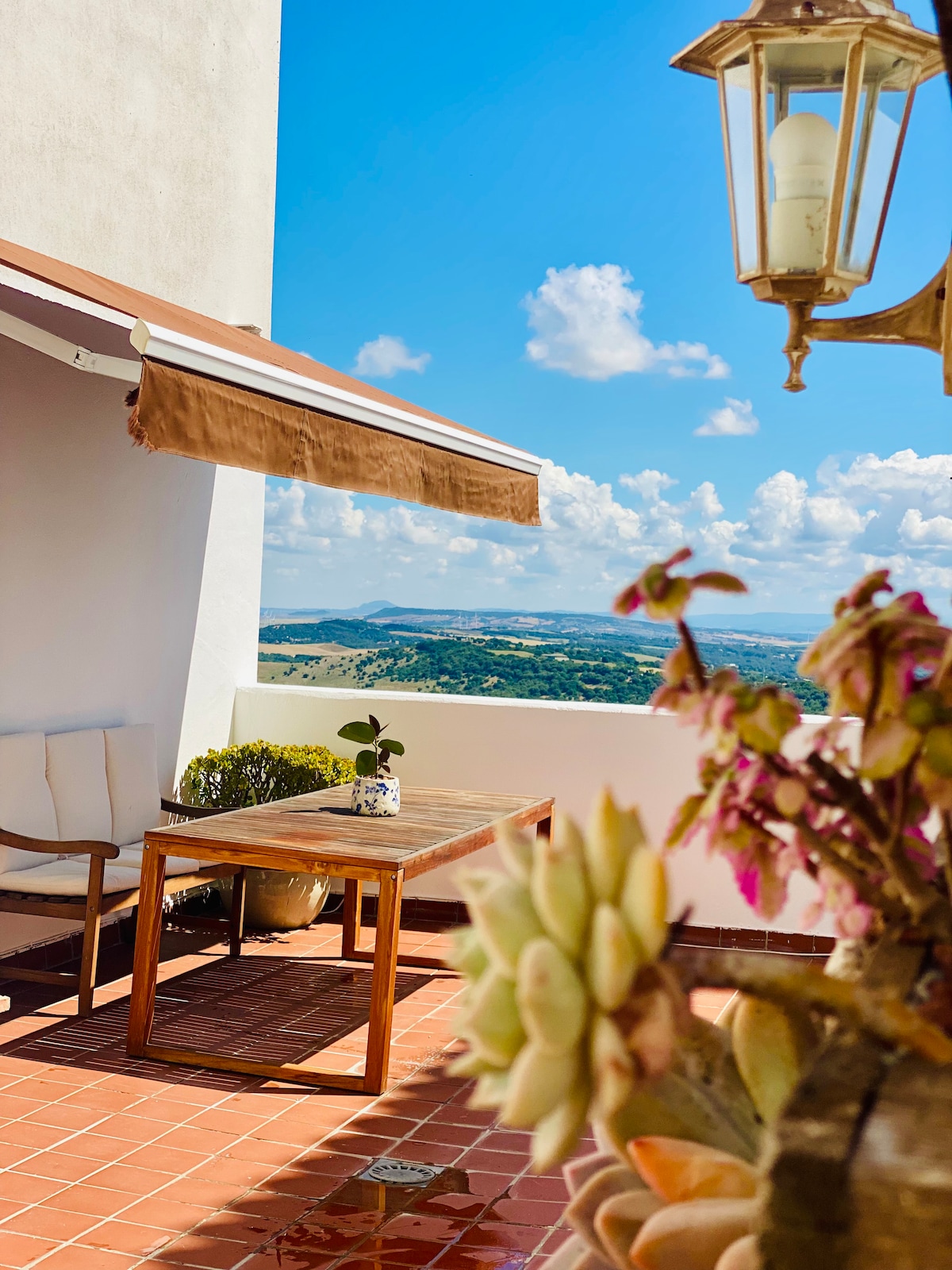 An inviting terrace showcases a wooden table surrounded by two chairs, offering panoramic views of the landscape. Brightly colored plants add vibrant touches, while a decorative lamp hangs overhead, enhancing the serene outdoor space. A clear blue sky complements the scenic backdrop.