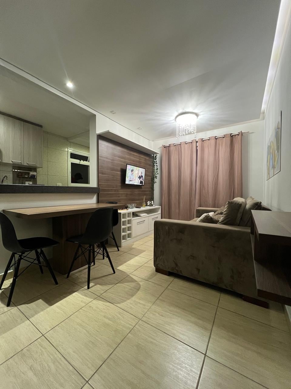 A compact living space features a small dining table with two black chairs adjacent to a comfortable sofa. A wooden console is positioned against the wall, and natural light filters through long, light brown curtains. The kitchen area is visible in the background.