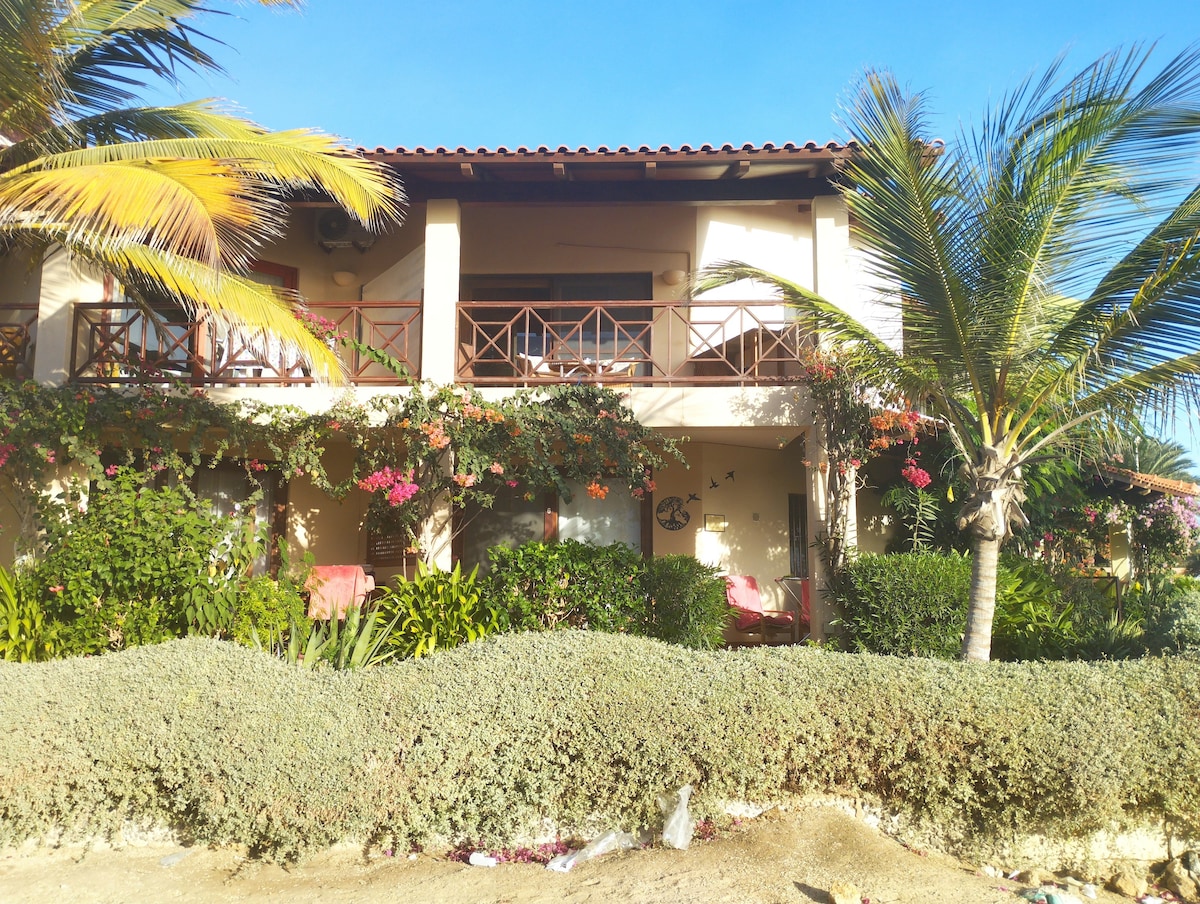 The residence features a two-story exterior with a balcony adorned by wooden railings. Lush greenery and flowering plants frame the entrance, while palm trees provide shade. The building's architecture reflects a welcoming beachfront atmosphere.