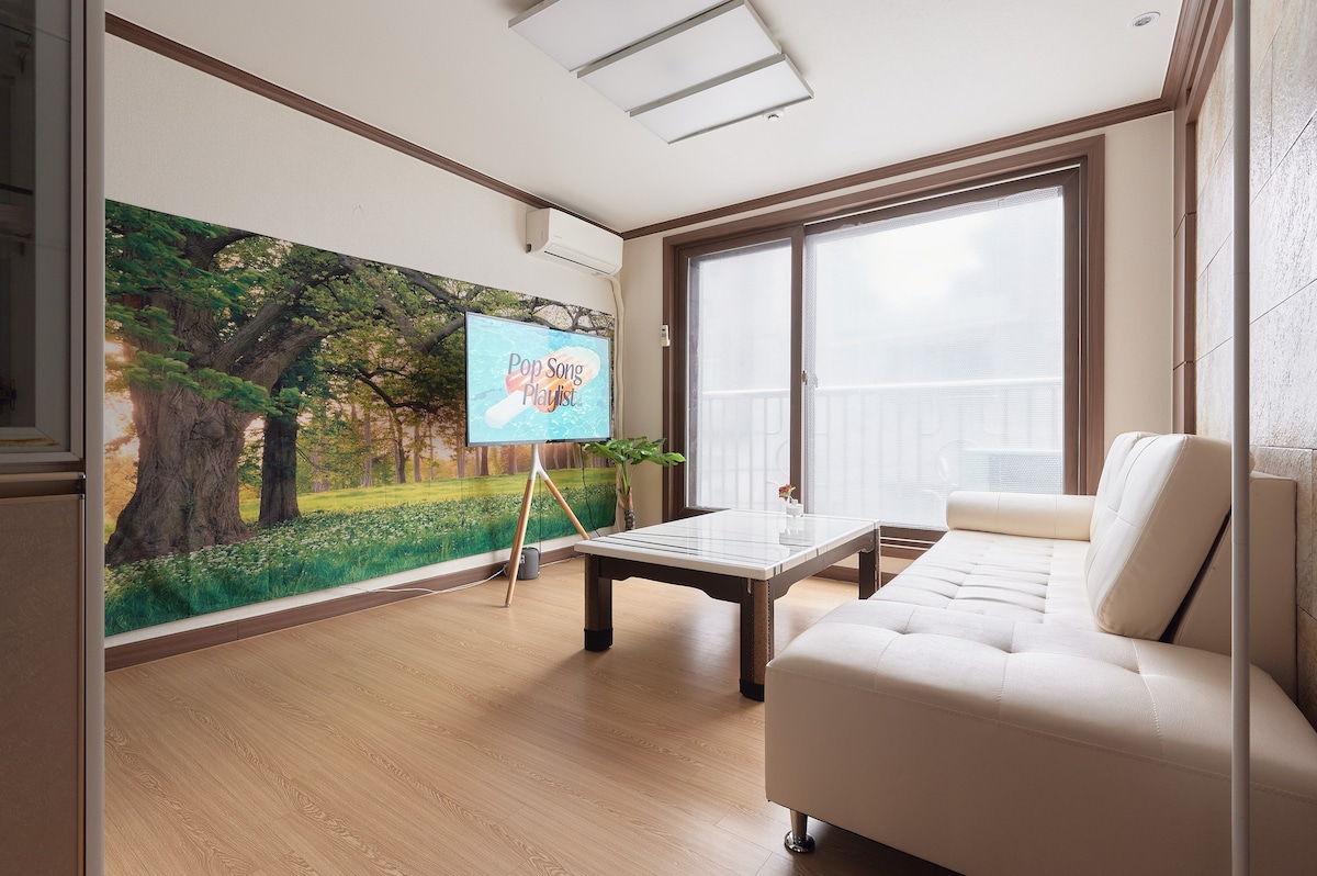 A bright living space features a minimalistic white sofa facing a wooden coffee table. A large window allows natural light to fill the room, complemented by a nature-themed wall mural. An air conditioning unit and a standing television on an easel enhance the functional layout.