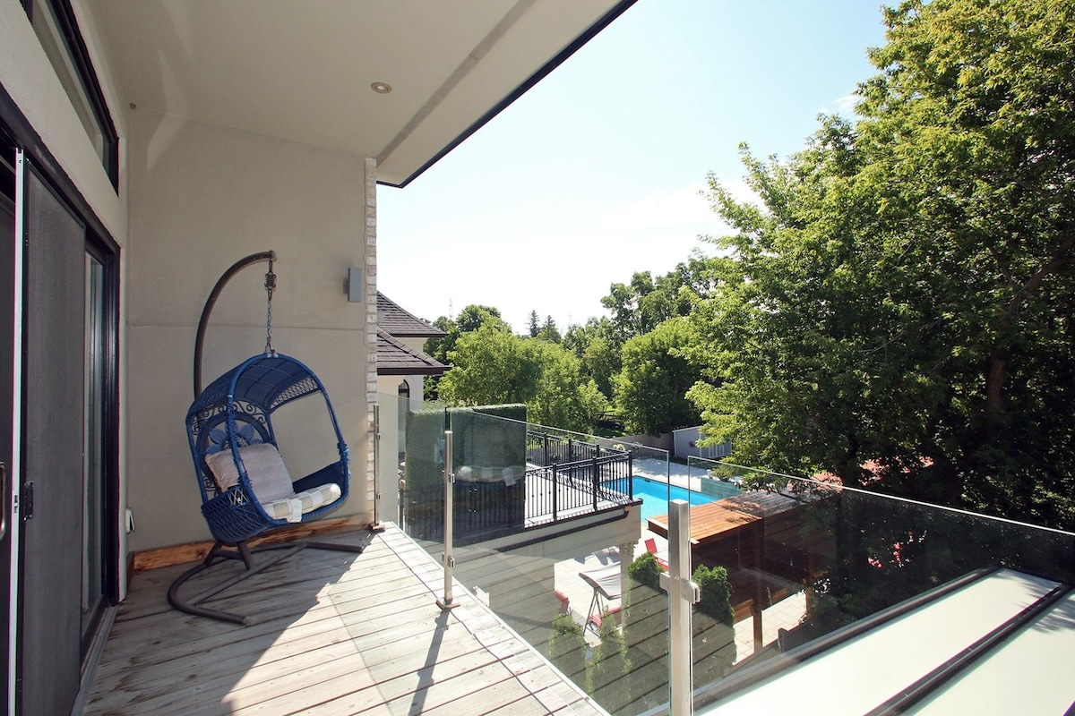 A covered balcony features a hanging blue chair with white cushions, providing a comfortable spot to relax. Glass railings enhance the view of the lush green trees and a pool area below, creating a serene outdoor setting.