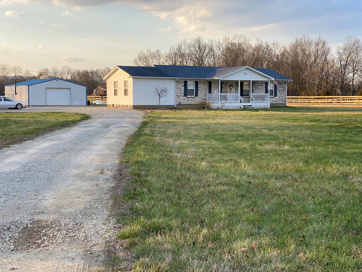 A single-story home is positioned on a sprawling green lawn, with a gravel drive leading to the entrance. The property features a covered front porch and is surrounded by trees and a fenced area. A spacious outbuilding is visible in the background.