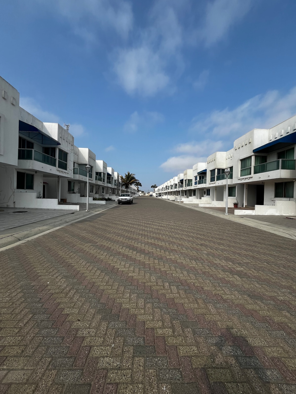 A wide, paved street is lined with modern, white buildings featuring large windows. The scene is brightened by a blue sky with scattered clouds, and palm trees are visible in the distance, contributing to a relaxed, coastal atmosphere.
