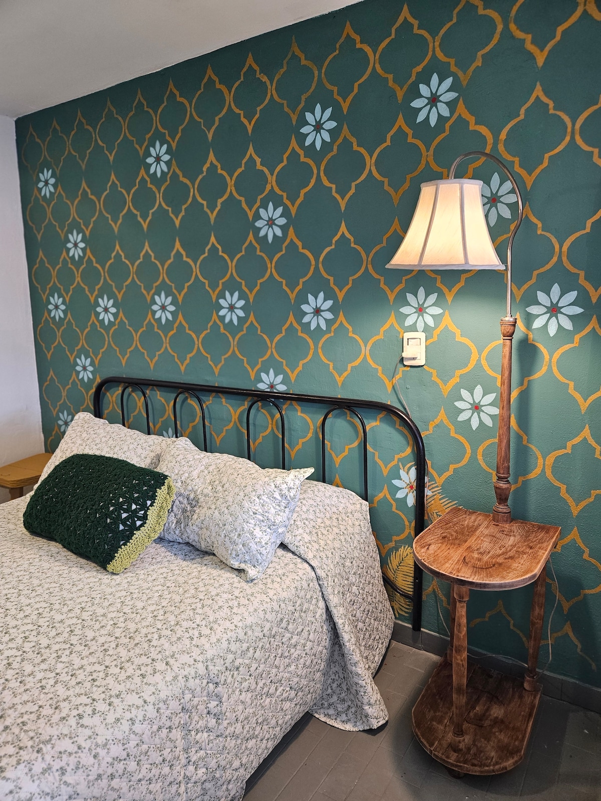 A sleeping area features a decorative wall in deep green with floral patterns. A patterned bedspread drapes over a spacious bed, complemented by a knitted green pillow. A wooden bedside table holds a lamp with a soft shade, providing a subtle source of light.