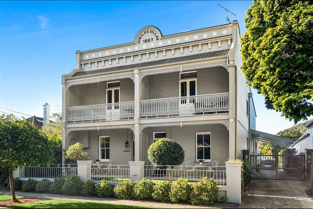 A charming two-story building built in 1887 features a classic façade with white painted brick. Two spacious balconies are adorned with decorative railings, and large windows allow natural light to fill the interior. Lush greenery surrounds the property, enhancing its inviting presence.