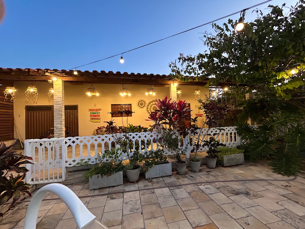 An outdoor area features a well-maintained patio, surrounded by various plants in decorative pots. Soft lighting hangs above, creating a relaxing ambiance. The building, with a tiled roof, is set against a twilight sky, offering a serene atmosphere for family gatherings.