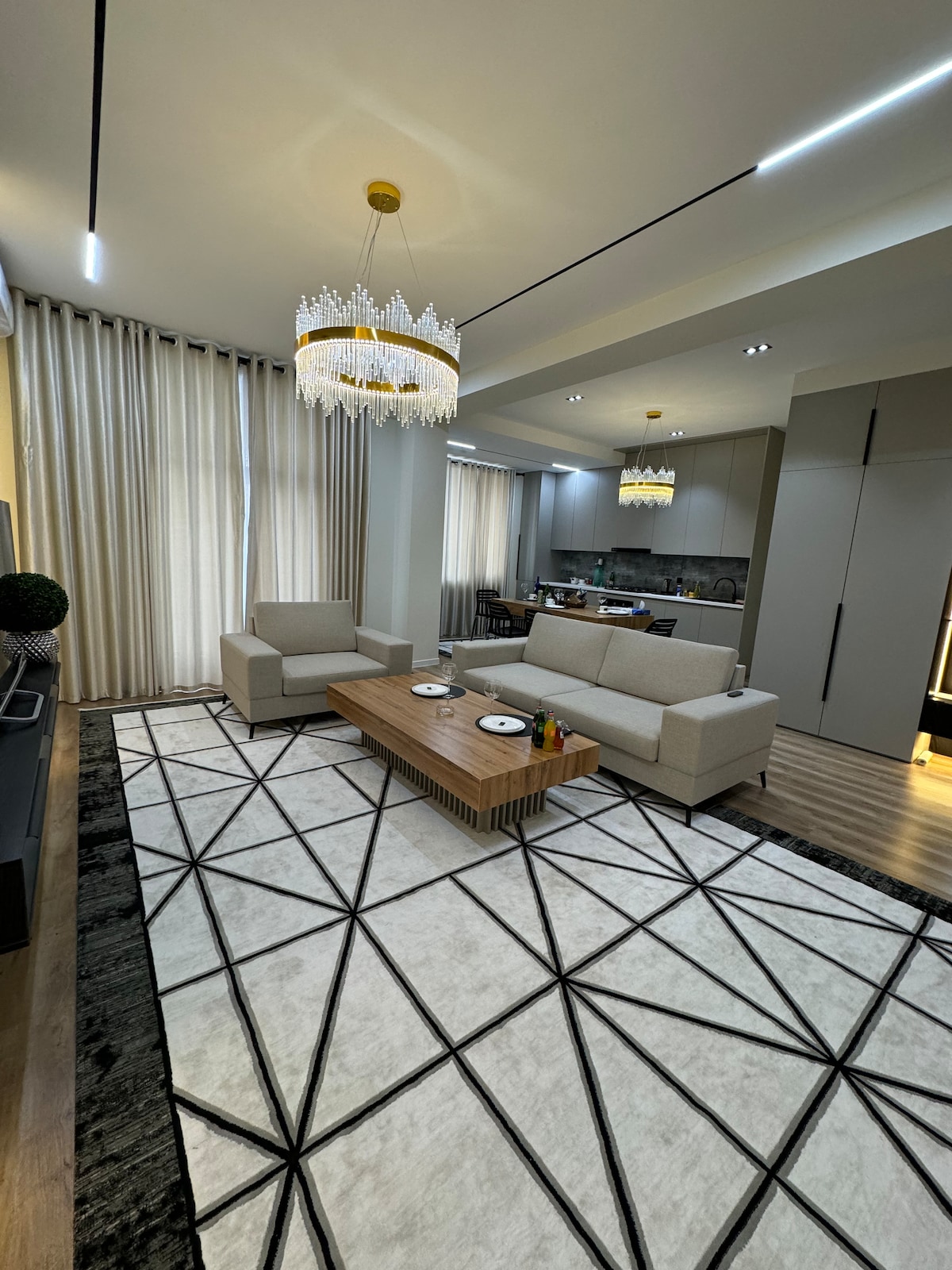 A spacious living area is presented with two light-colored sofas positioned around a wooden coffee table. A stylish chandelier hangs from the ceiling. Large windows dressed with drapes allow natural light to fill the room, which features a geometric patterned rug.