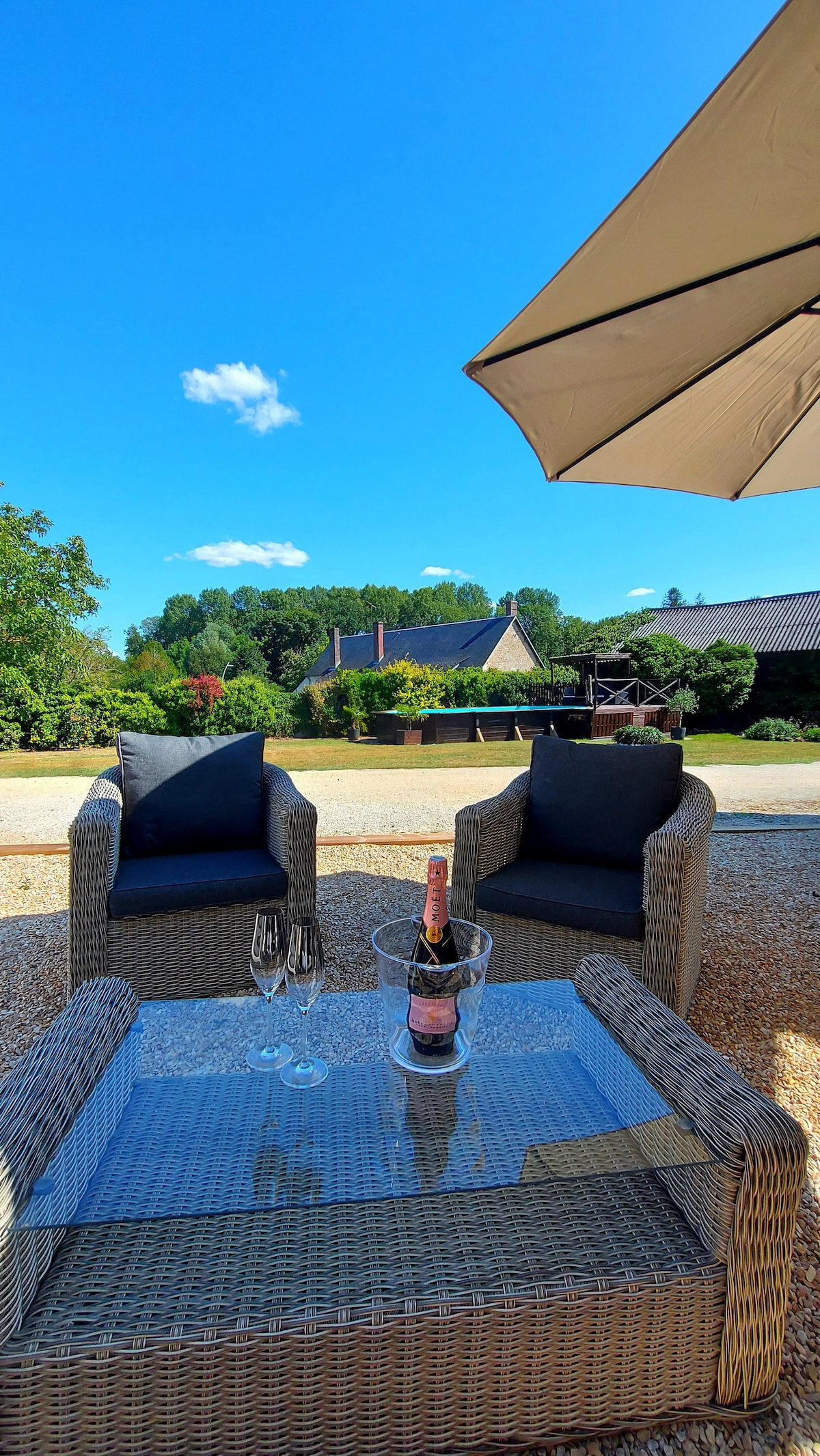 An outdoor seating area is presented with two comfortable chairs and a glass-top table. A bottle of champagne is accompanied by two glasses. A large umbrella provides shade while a scenic garden and distant structures fill the background under a clear blue sky.