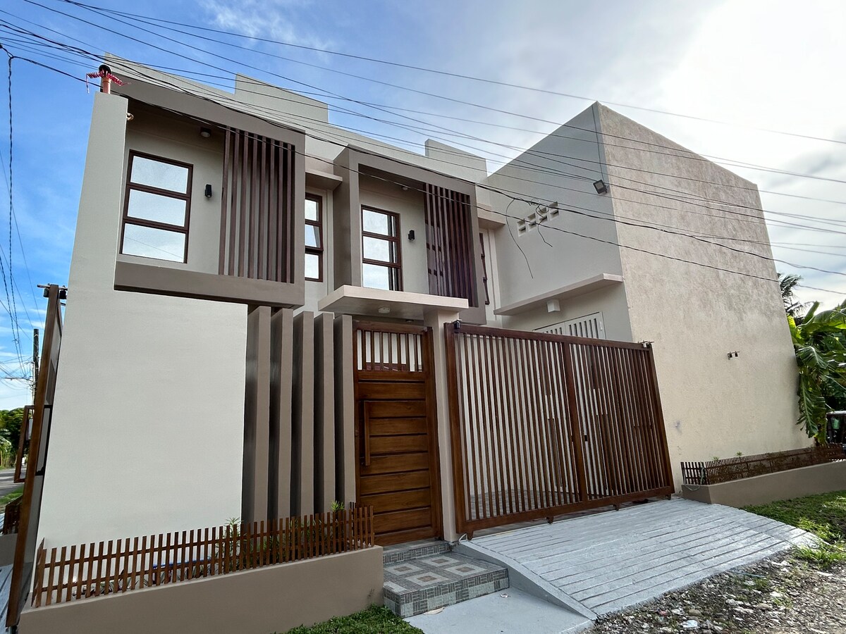 A modern two-story building features a combination of light and dark exterior walls, with wooden slat accents. A wooden gate provides access, and a paved pathway leads to the entrance. The structure is complemented by a mix of large windows, allowing natural light to enter.