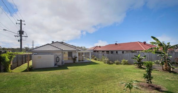 A spacious backyard is presented with lush green grass, bordered by a wooden fence. A single-story house is visible, featuring a covered patio area. Nearby, several banana trees and shrubs add to the garden's natural ambiance under a clear blue sky.