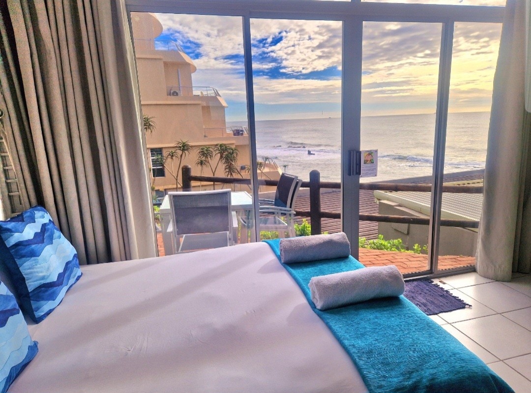 A spacious studio features a large bed adorned with a light blue throw and two white towels. Floor-to-ceiling windows provide a view of the beach and sky. Outdoor seating is visible on the balcony, with gentle waves lapping at the shore in the background.
