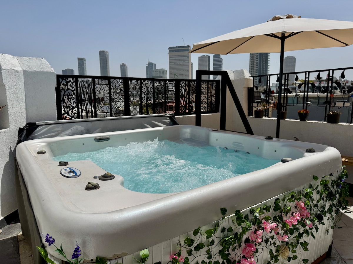 A private jacuzzi is situated on the spacious terrace, surrounded by colorful flowers. Umbrellas provide shade, enhancing comfort while enjoying the views of the skyline. The bubbling water of the jacuzzi creates an inviting atmosphere for relaxation.