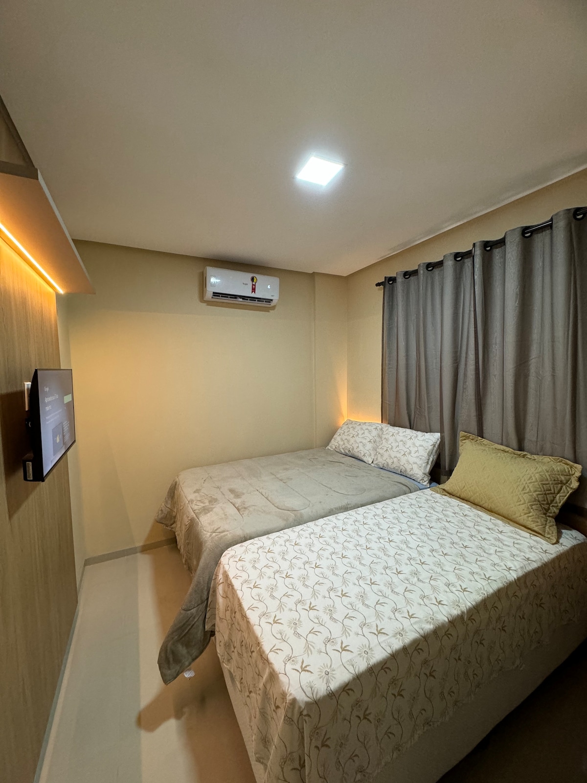 A well-appointed bedroom is depicted, featuring two beds with decorative bedding and pillows. A flat-screen television is mounted on the wall opposite the beds. The space is illuminated by overhead lighting and has soft curtains covering the window.