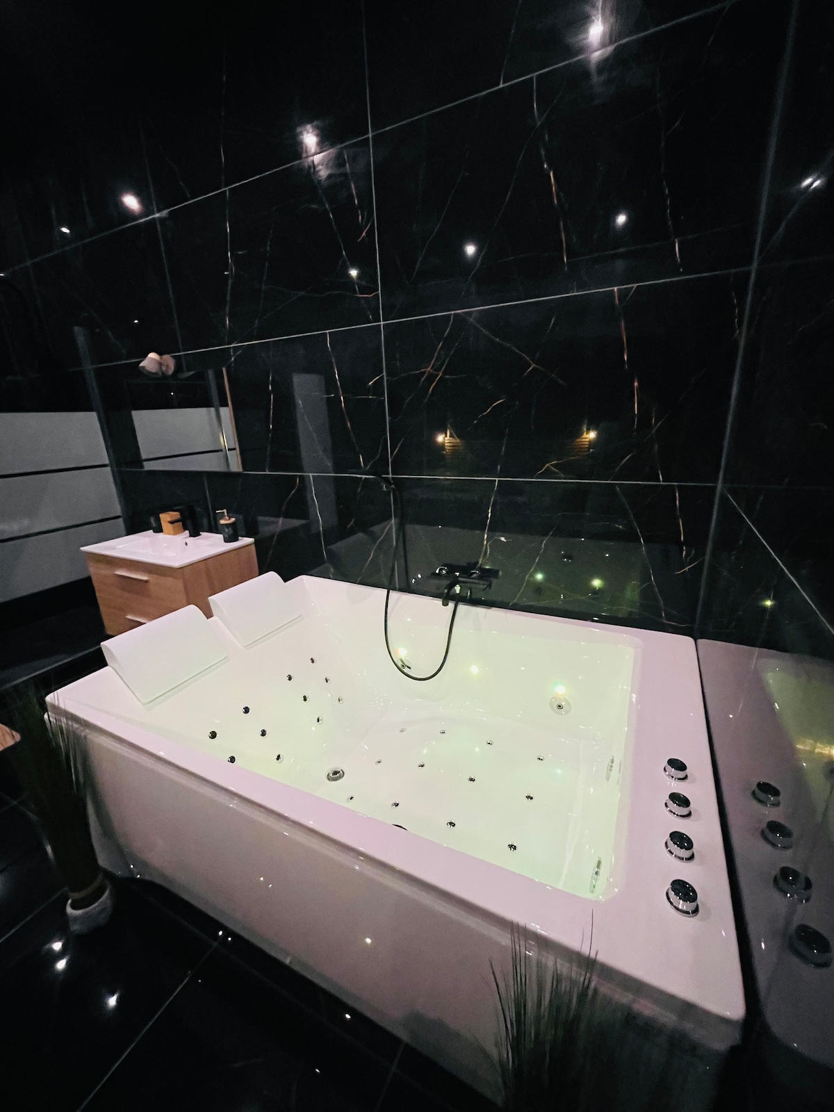 A private whirlpool bath is featured, surrounded by sleek black tiles that reflect soft lighting. The spacious tub includes multiple jets along the sides, creating a luxurious and relaxing atmosphere. A minimalist wooden cabinet is positioned nearby, enhancing the modern design.