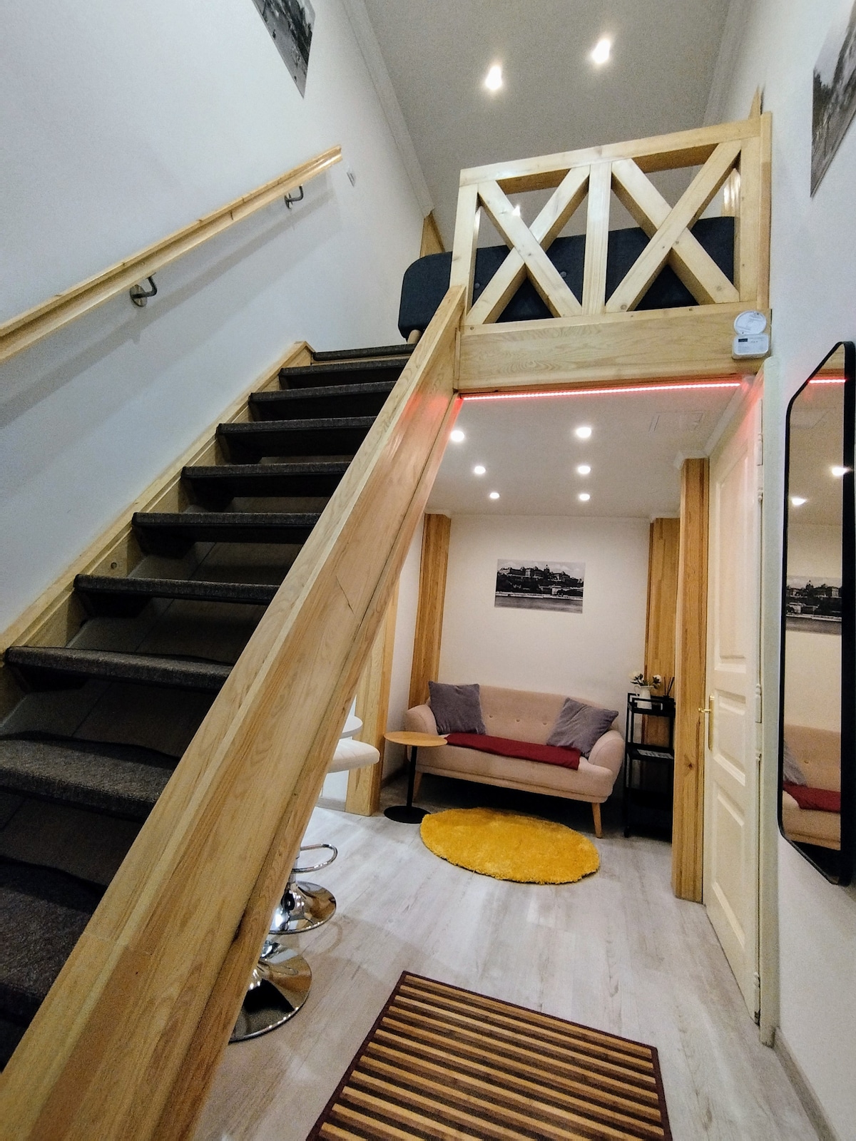 A staircase with wooden steps leads to a loft area, with a cozy seating area visible below. The entryway is well-lit, featuring a round yellow rug and a mirror reflecting the space. Soft seating is arranged for comfort and relaxation.