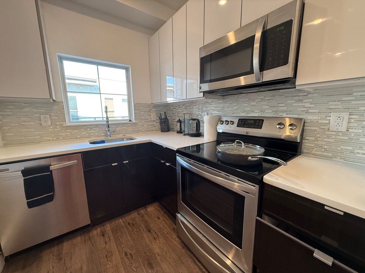The kitchen is designed with modern cabinetry featuring a mix of dark and light finishes. Stainless steel appliances, including an oven and microwave, are present. A window allows natural light to fill the space, highlighting the clean lines of the quartz countertops and the stylish glass tile backsplash.